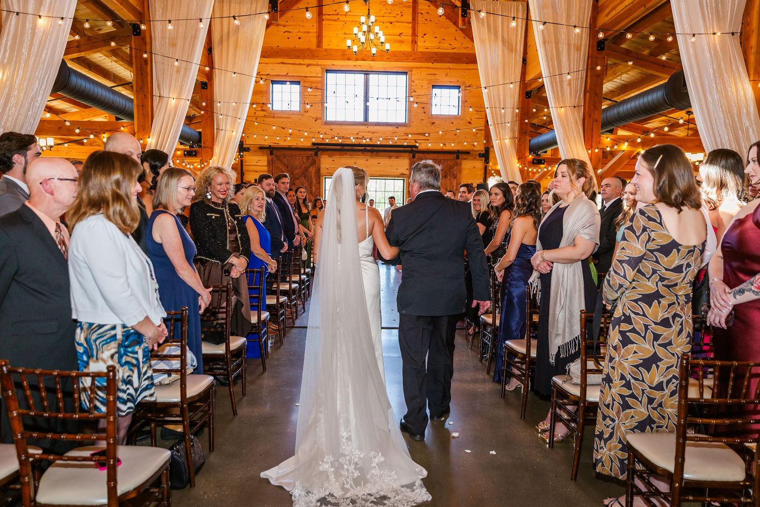 Elegant barn wedding ceremony with draped fabric, chandelier lighting, wooden beams, and guests seated along a floral aisle.