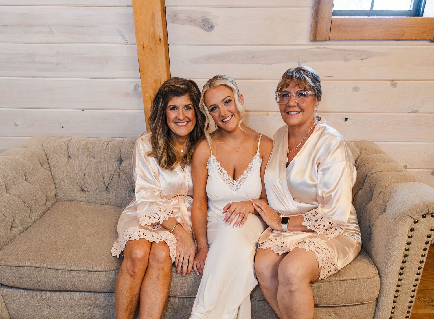 Three women in white satin robes and bridal gown smiling together on a beige tufted sofa in a rustic wood venue.