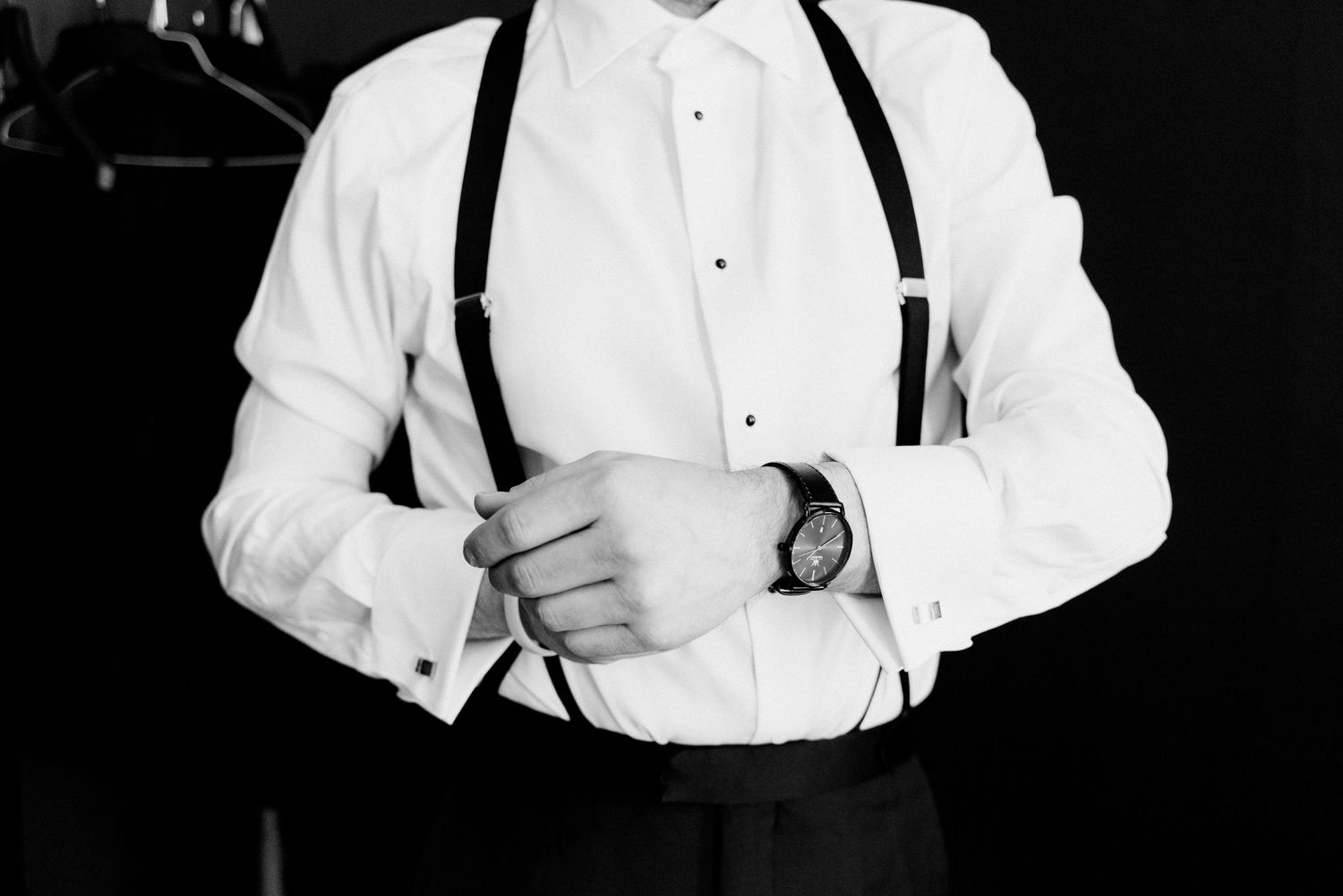 Black and white detail photo of a groom in white shirt and suspenders adjusting his cufflinks on wedding day.