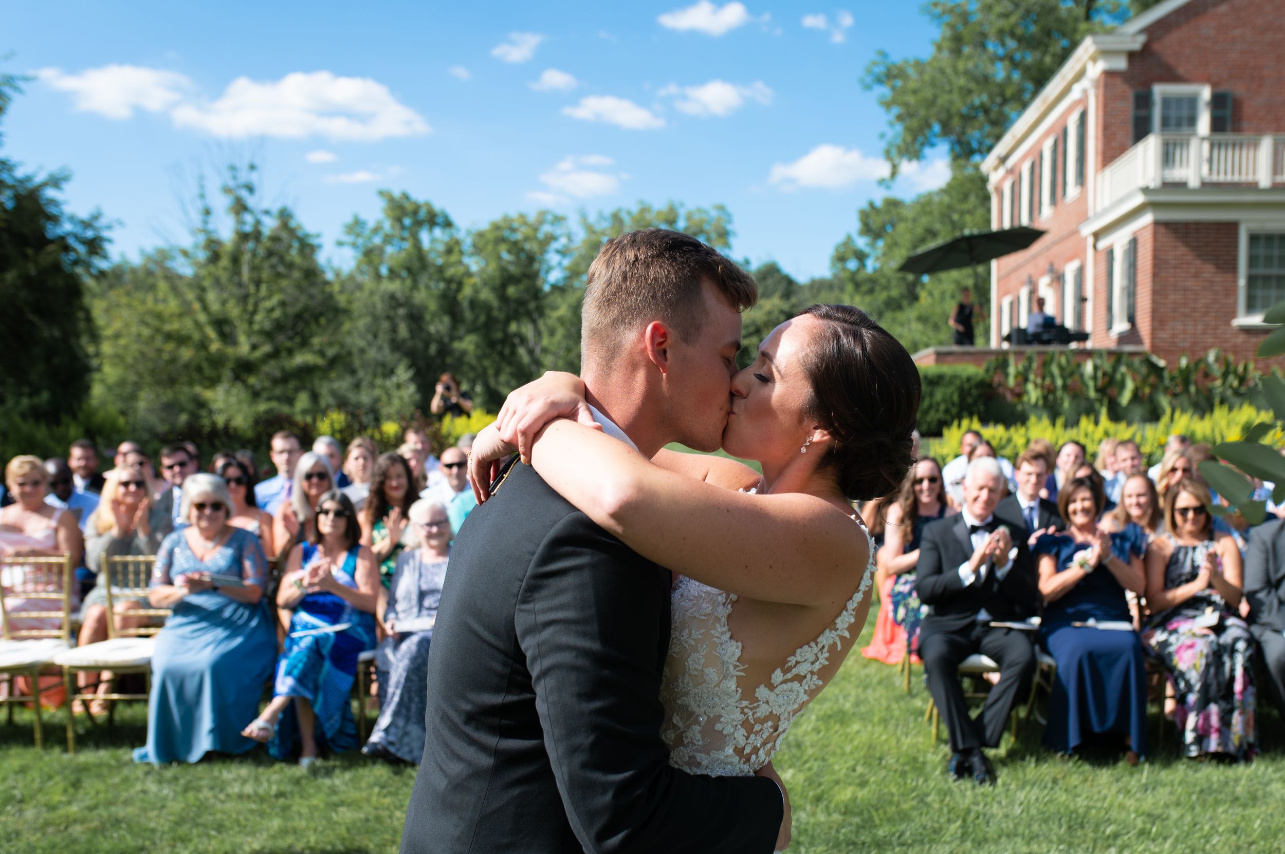 Bride and groom share first kiss at outdoor wedding ceremony with guests seated in gold chairs.
