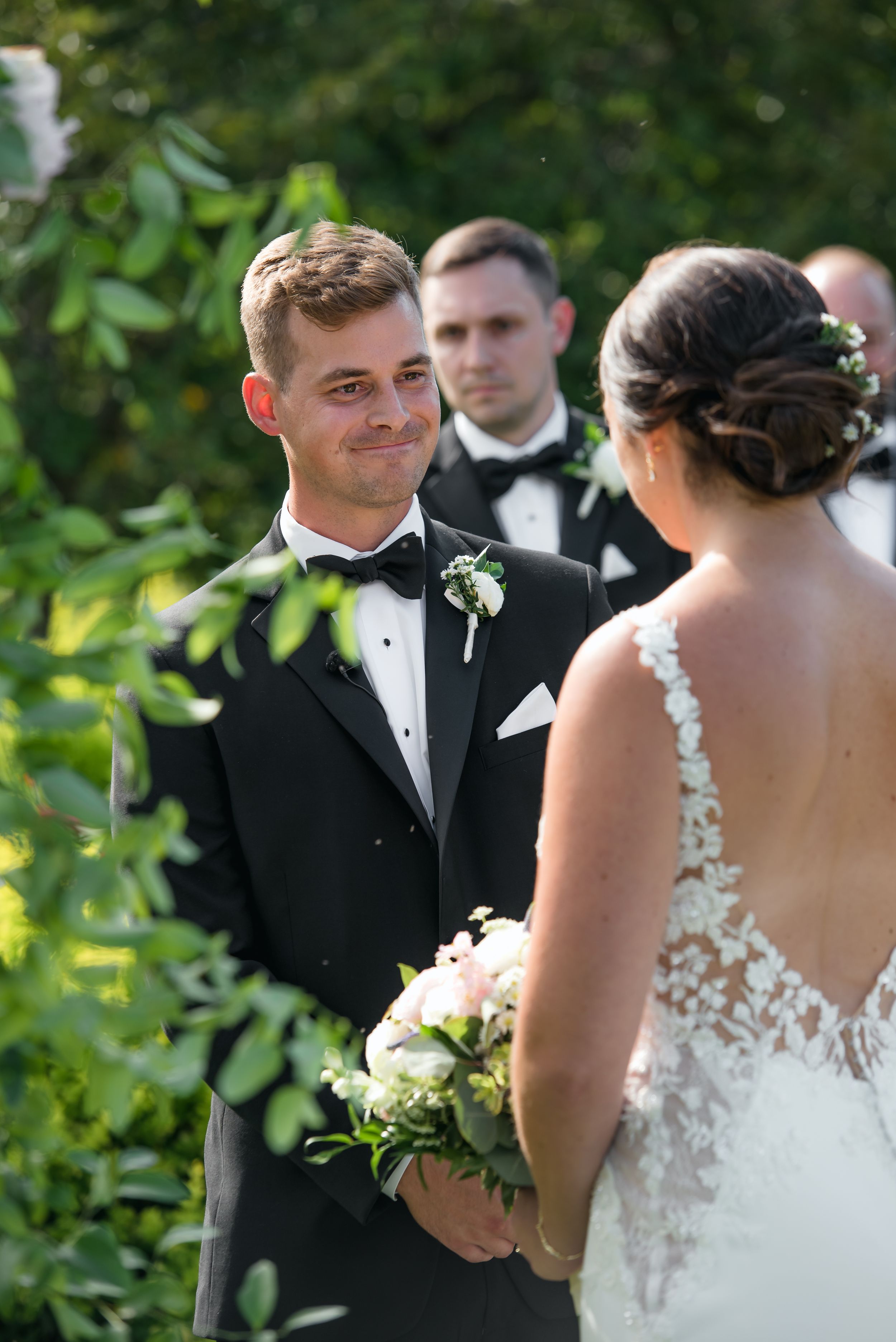 Groom in black tuxedo smiling emotionally at bride in lace wedding dress during outdoor ceremony.