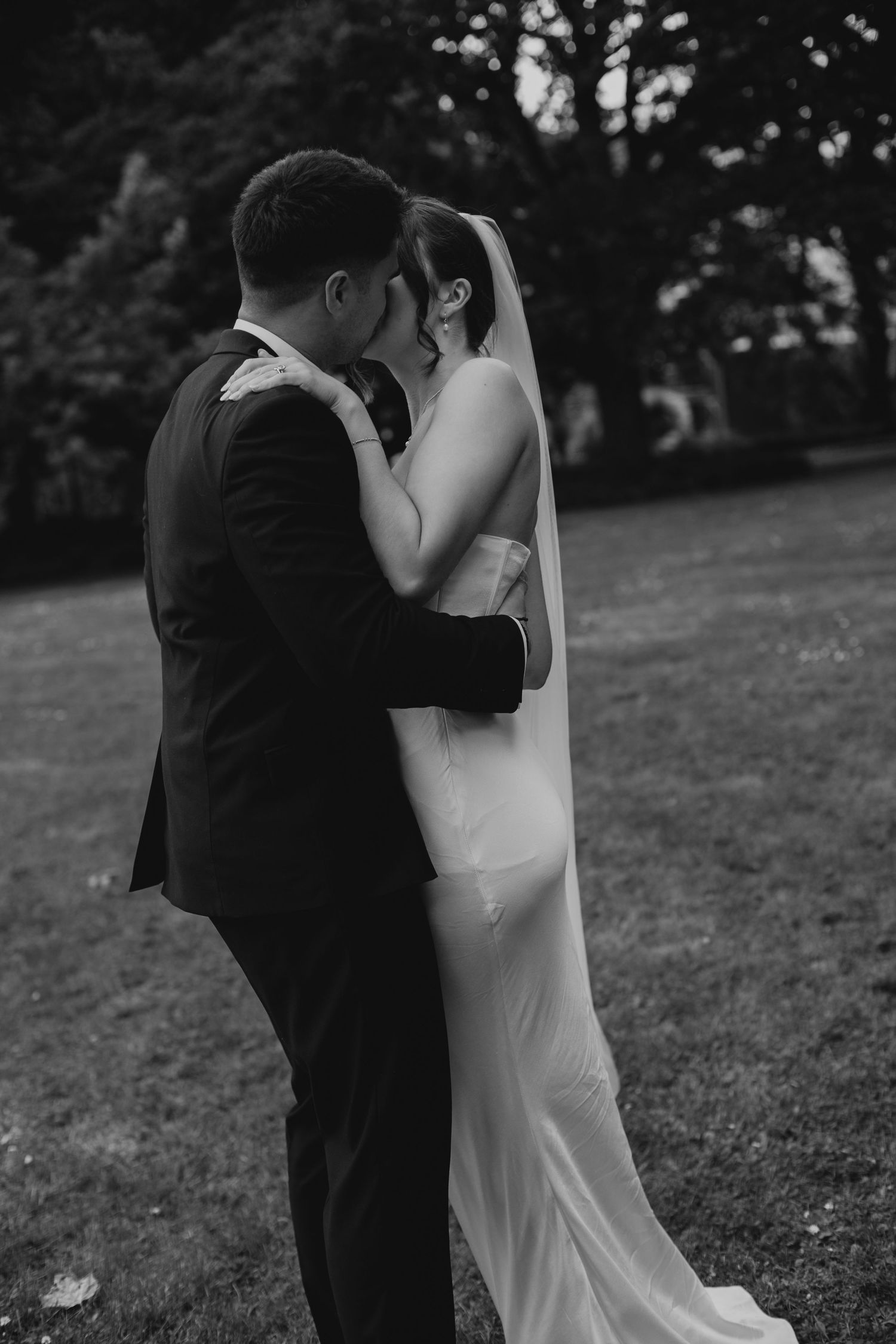 A bride and groom share a romantic kiss outdoors on their wedding day. The image is black and white.