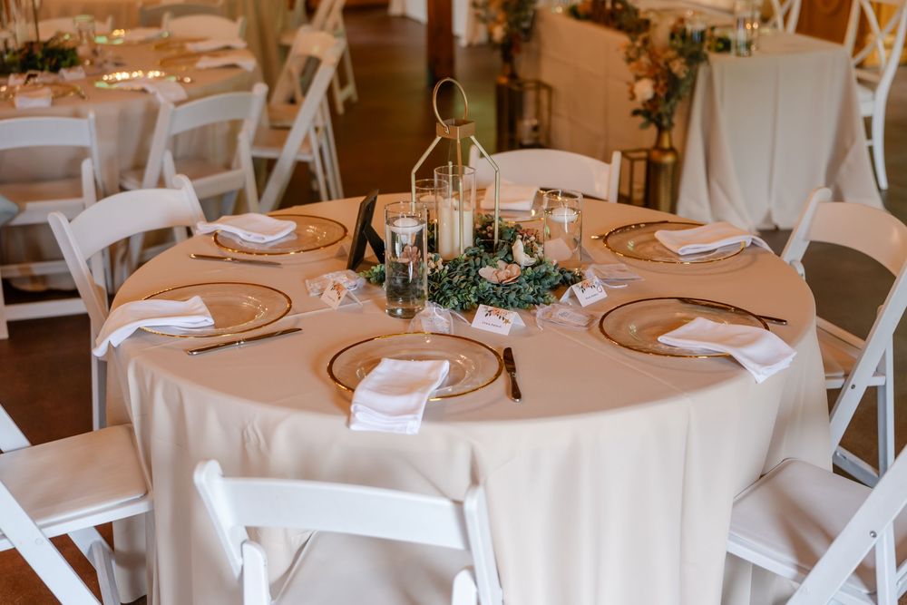 Elegant wedding reception table with white chairs, gold charger plates, candles, and greenery centerpiece on a beige tablecloth.