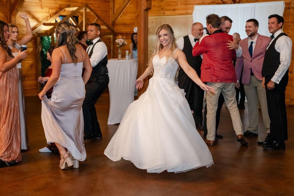 Bride in white ball gown dancing joyfully with guests at rustic wood-paneled wedding reception venue.