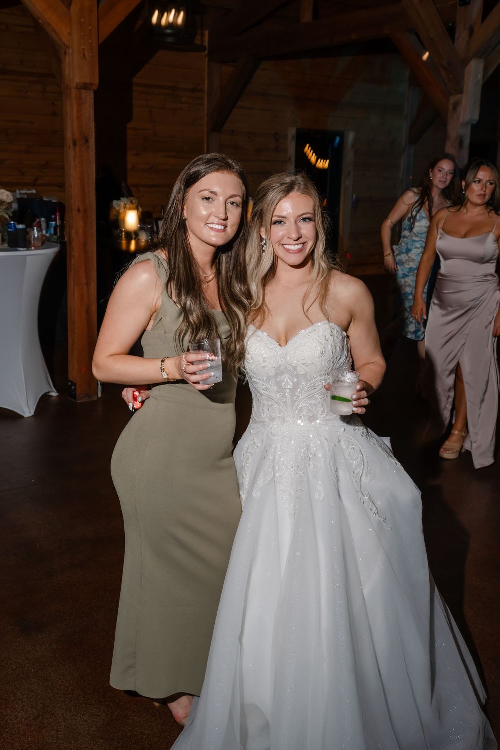 Bride in white lace gown poses with bridesmaid in sage green dress at rustic barn wedding reception.