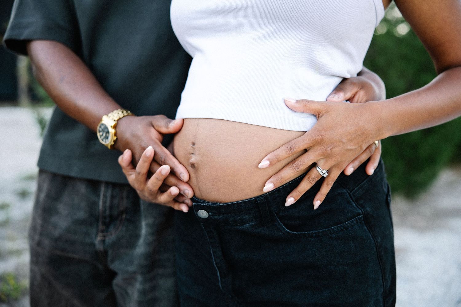 Couple holding pregnant belly together outdoors, showing maternity bump.