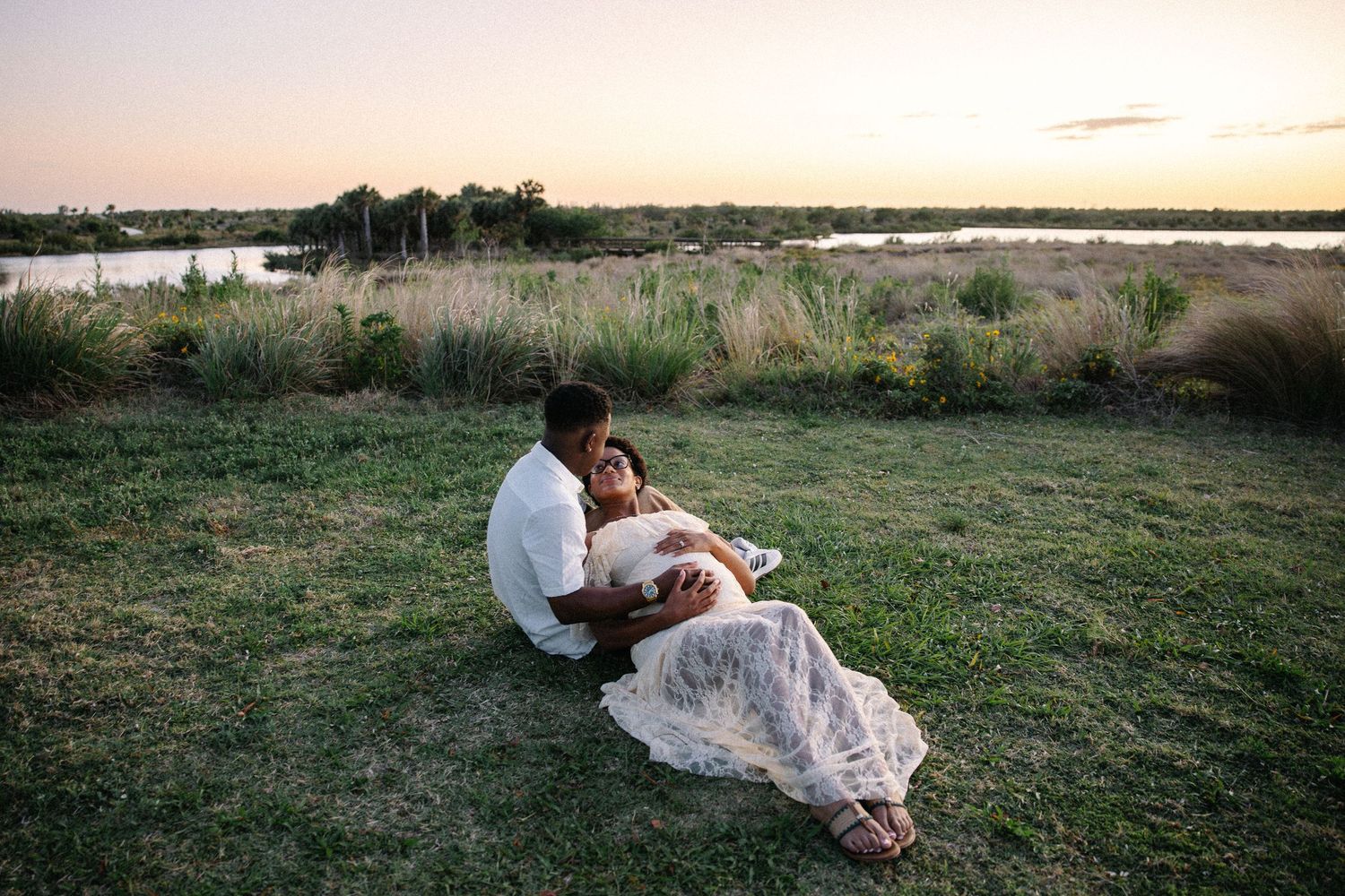 Couple sitting together on grassy riverbank watching a peaceful sunset.