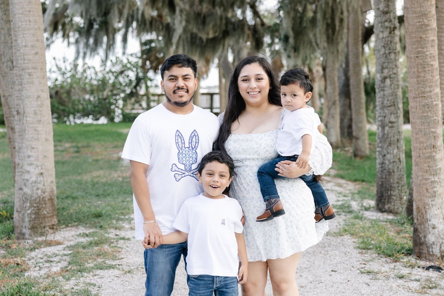 A happy family of four poses outdoors among palm trees on a sandy path, wearing coordinating white outfits.
