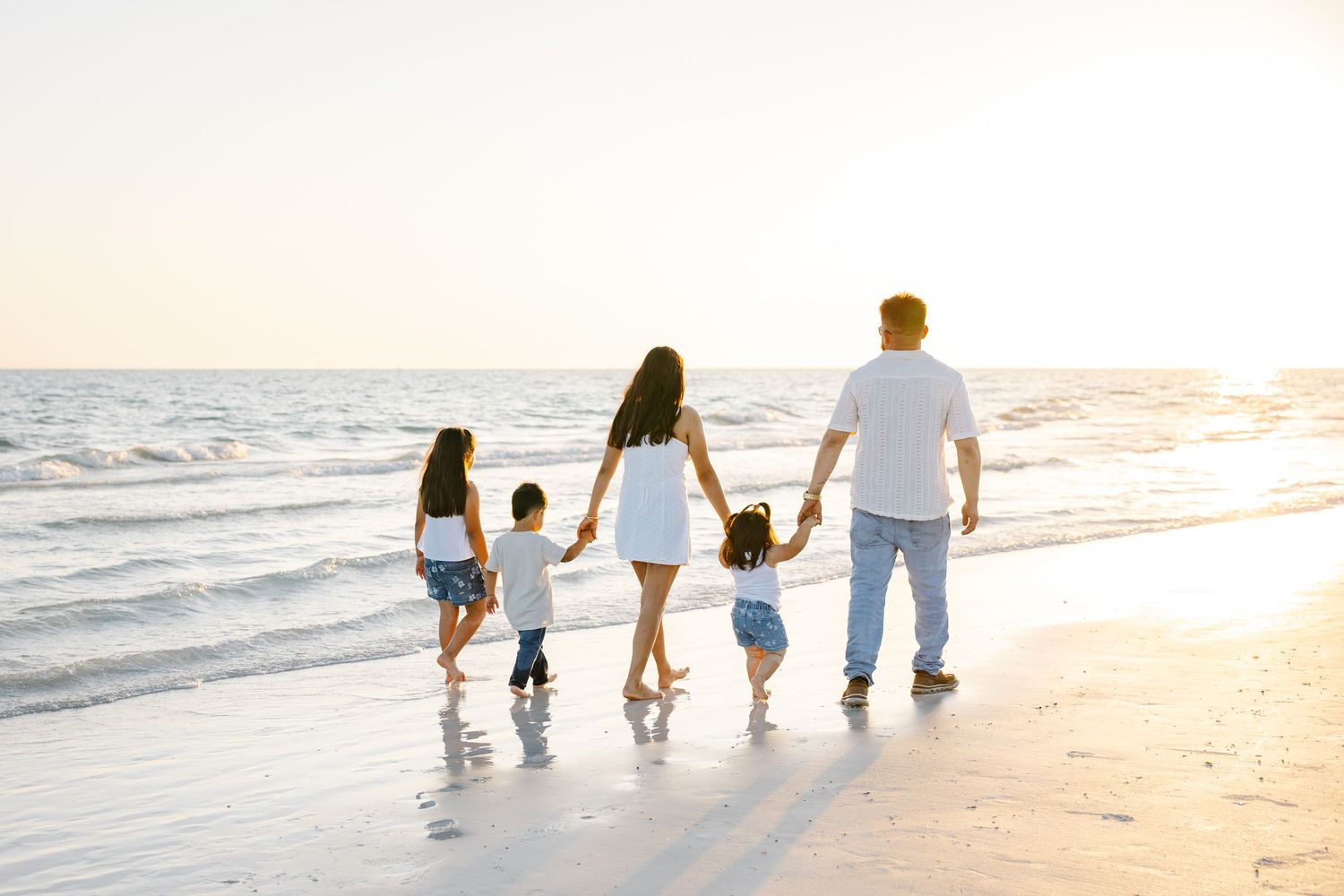 Family of five walks hand-in-hand along a sunlit beach at golden hour, viewed from behind.