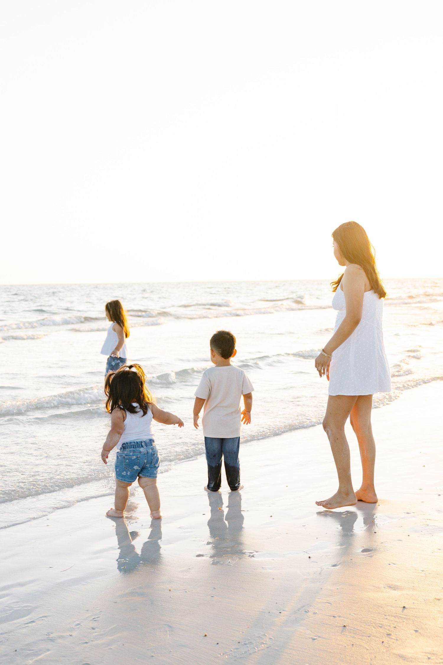 Mother watches her three young children run toward ocean waves on a bright sunny beach.