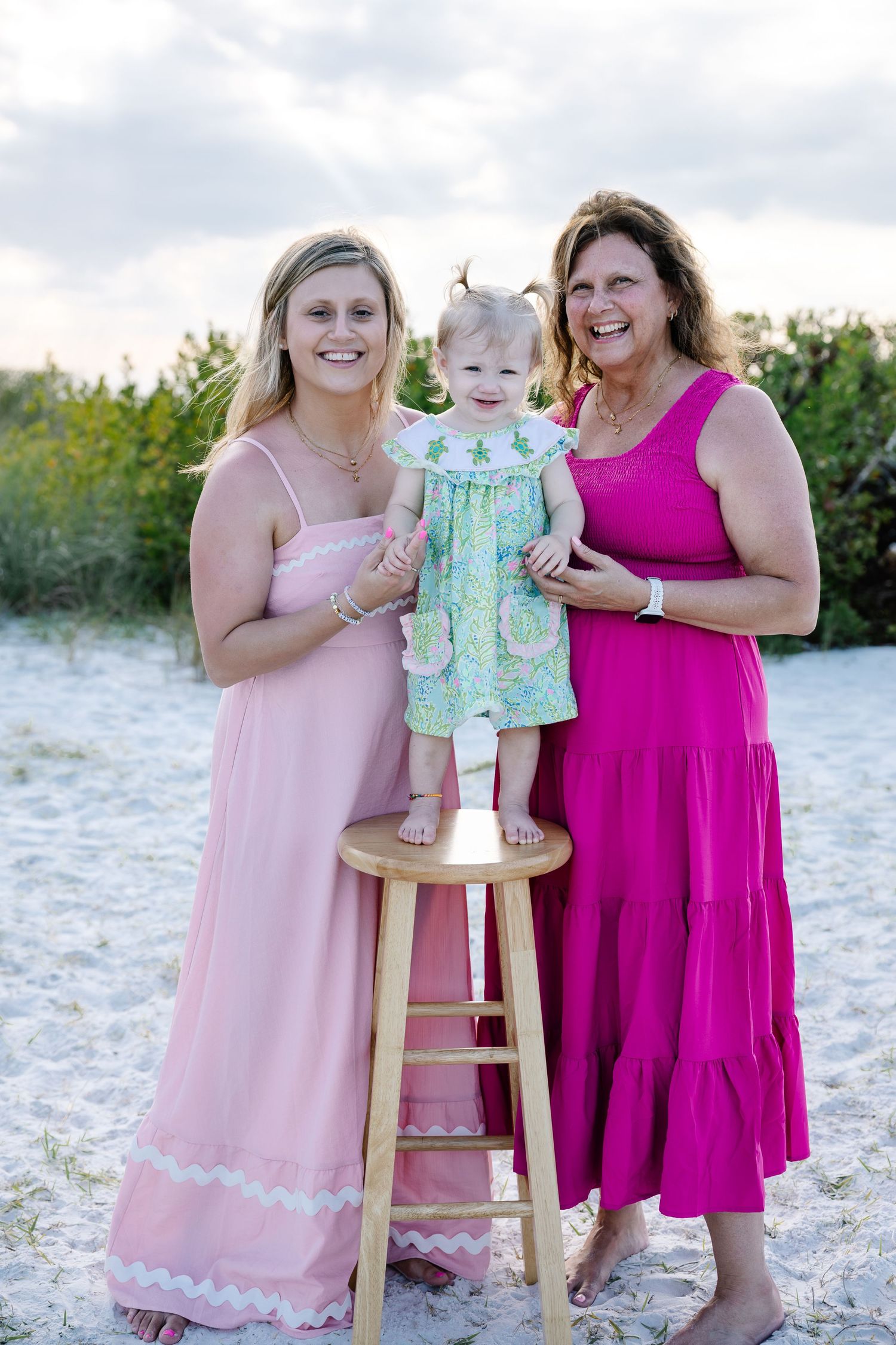 Three generations of women in pink dresses smile together on a sandy beach at sunset with a toddler.