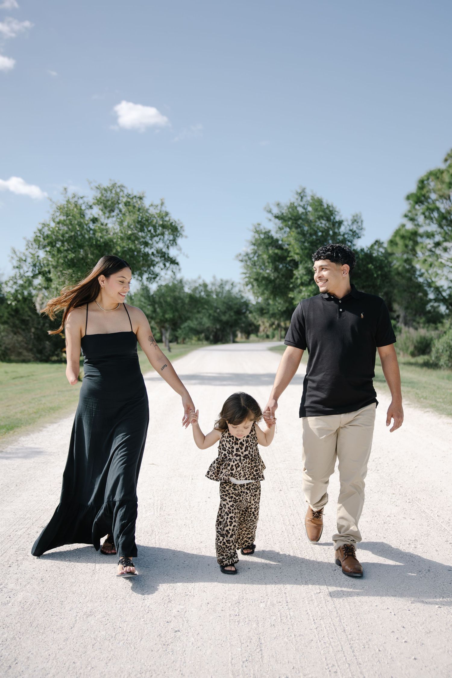 Happy young family holding hands and walking together down a sunny tree-lined gravel path outdoors.