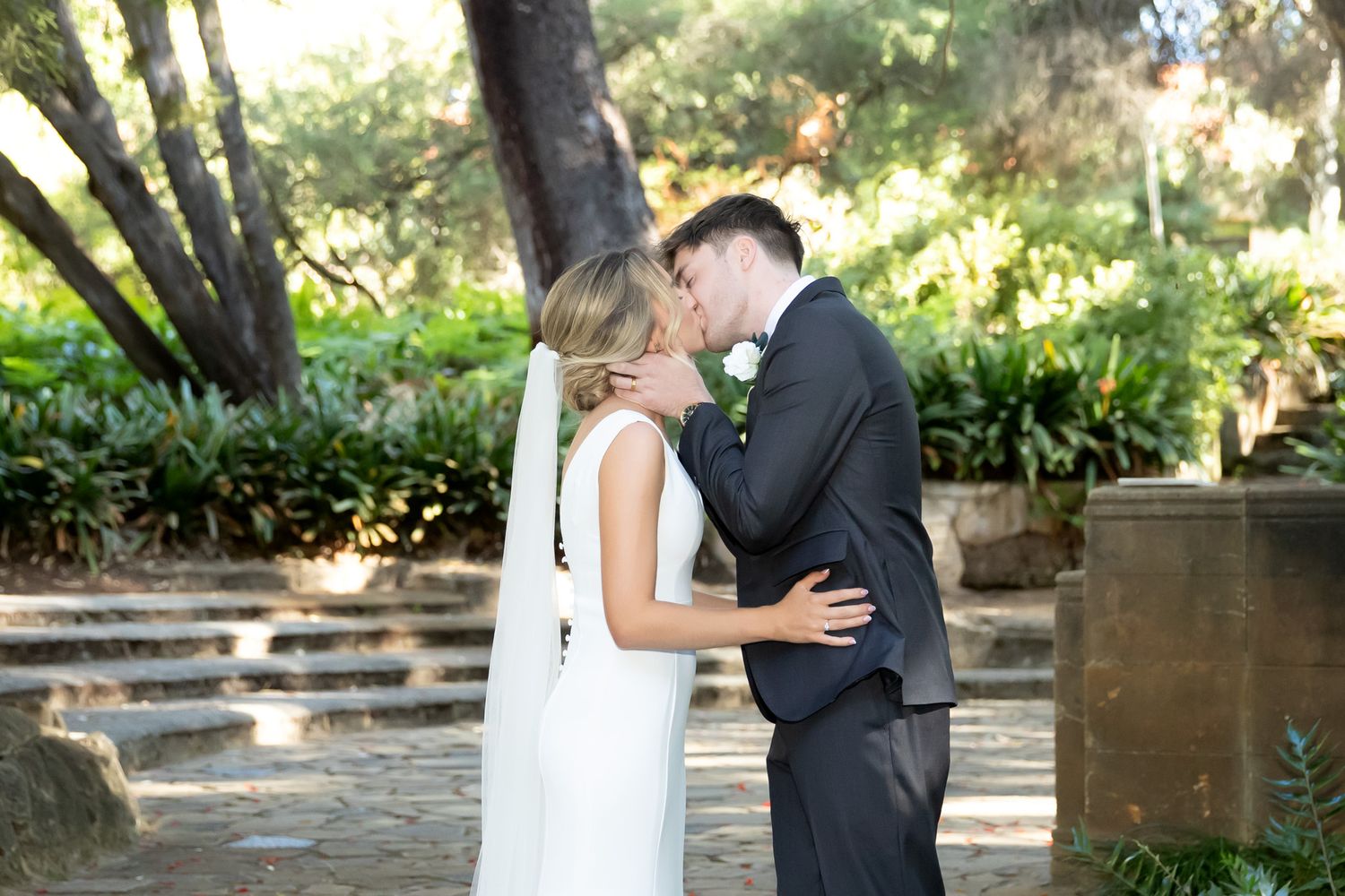 Bride and groom sharing a tender kiss surrounded by lush greenery in a sunlit outdoor garden.
