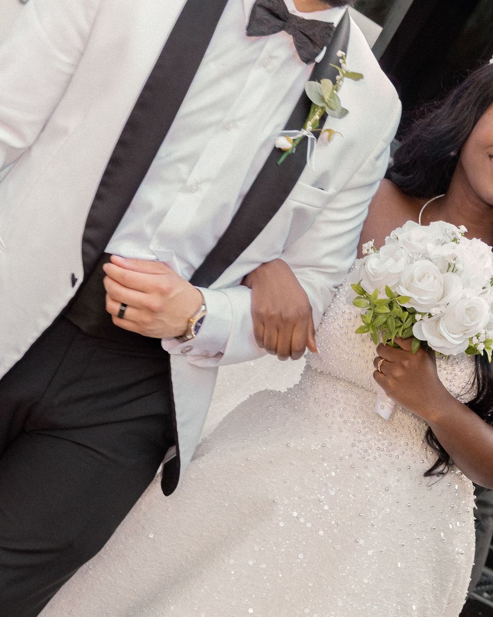 Bride in sparkly white gown holds white rose bouquet while linking arms with groom in white tuxedo jacket.