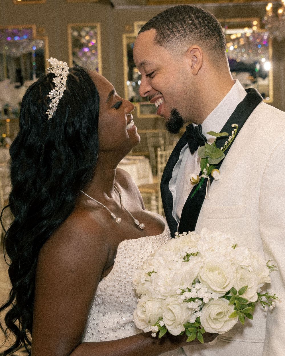 Smiling bride wearing tiara gazes up at laughing groom holding white rose bouquet in ornate ballroom.