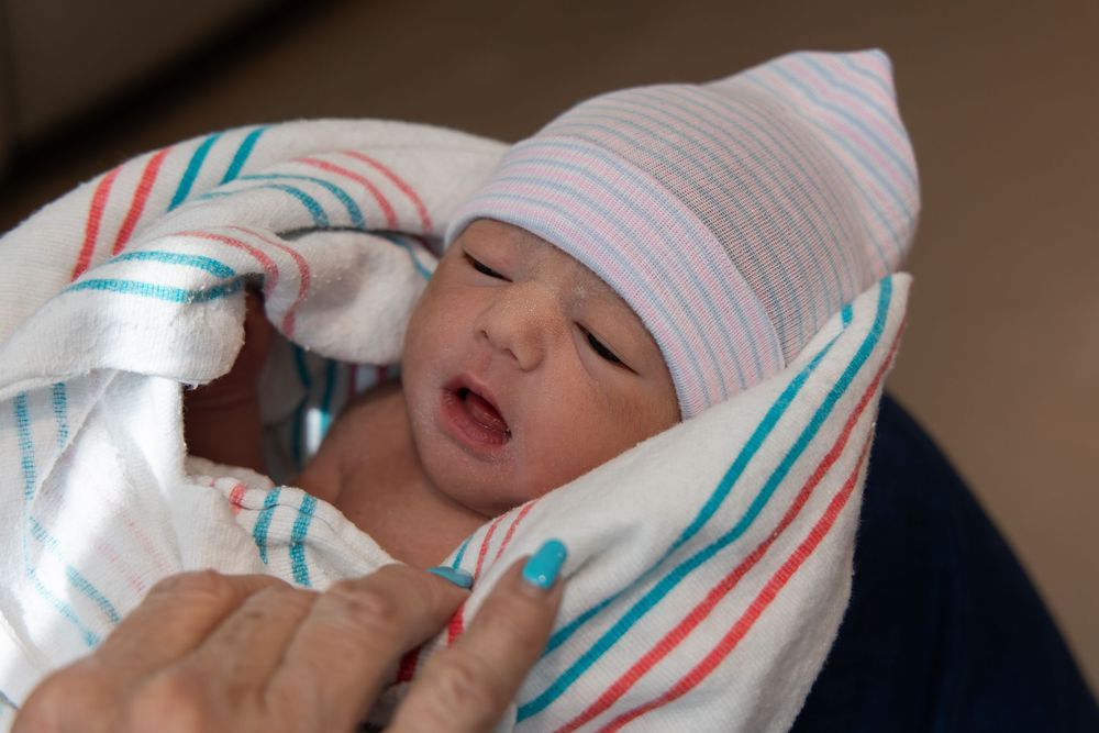 Newborn baby wrapped in striped blanket wearing white hospital hat, eyes closed, held by caregiver.