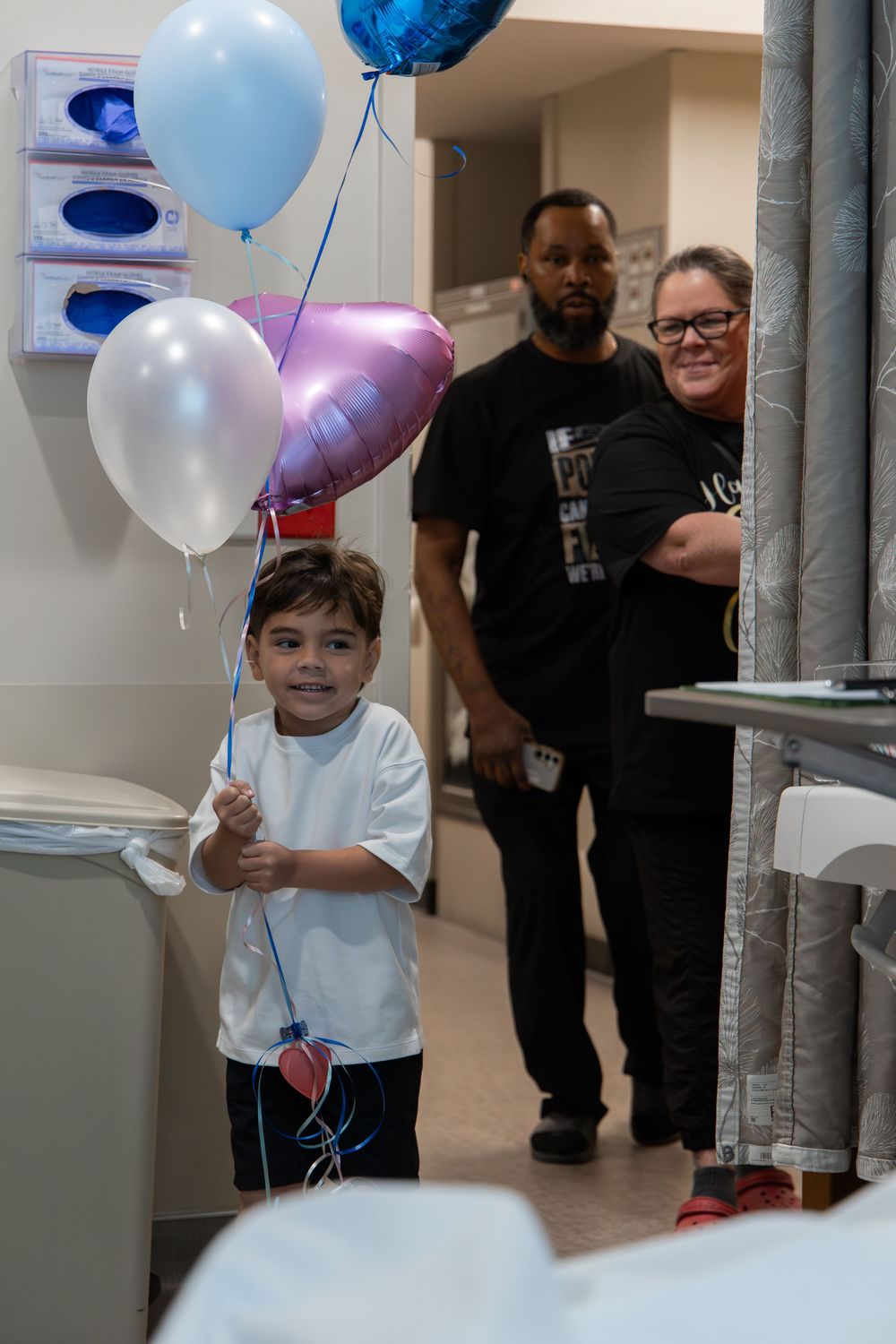 Young boy holding colorful balloons in a hospital room with two adults smiling in the background.