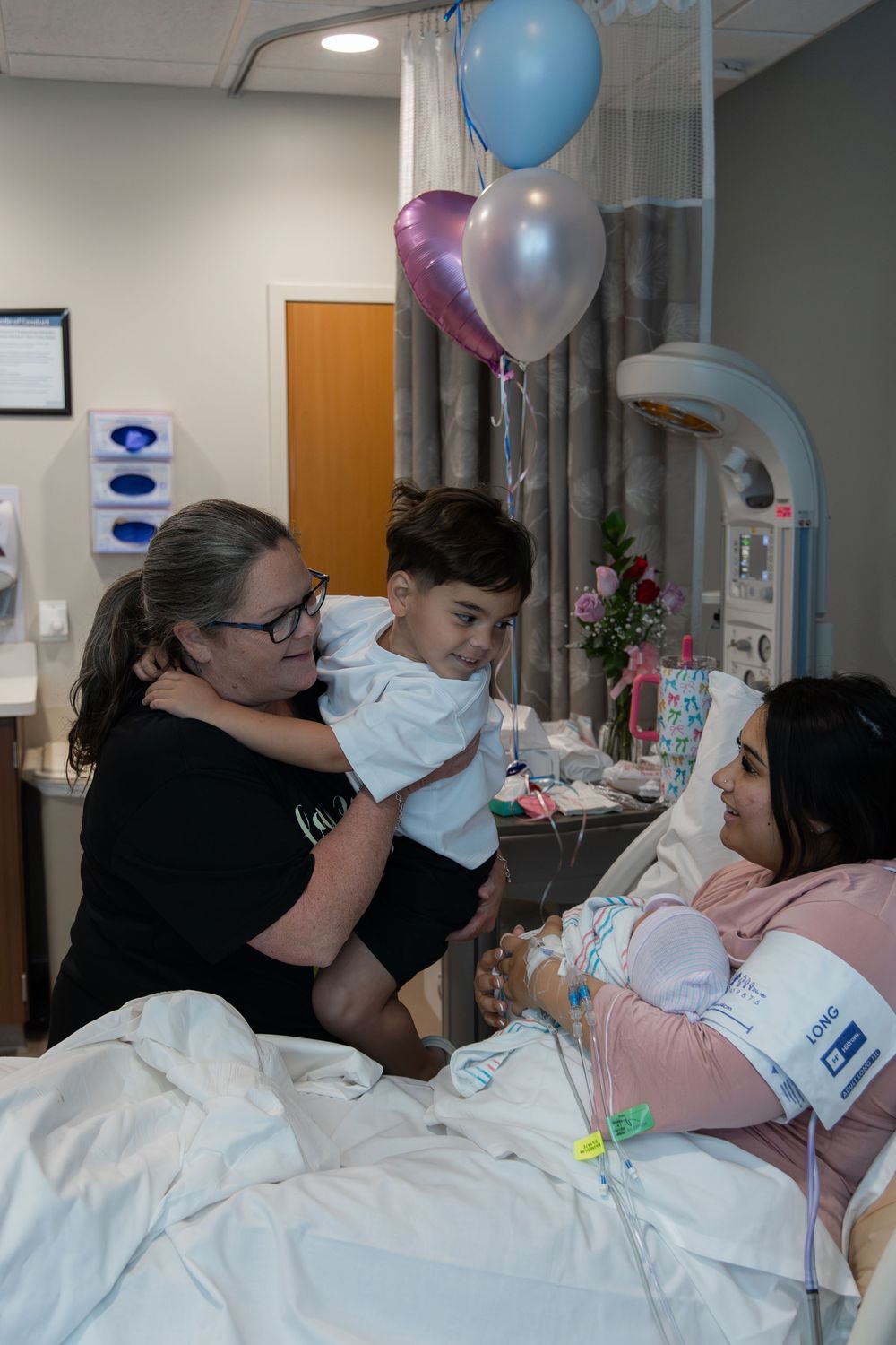 Woman holding toddler visits new mother in hospital bed, with pink and blue balloons celebrating a newborn.