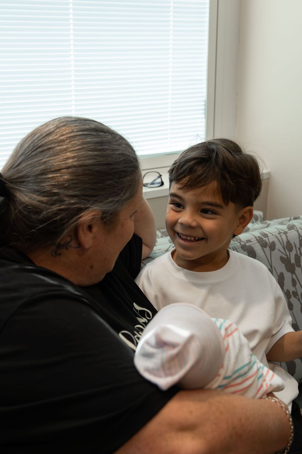 An adult holding a newborn baby smiles warmly at a young boy in a white shirt, sharing a tender family moment indoors.