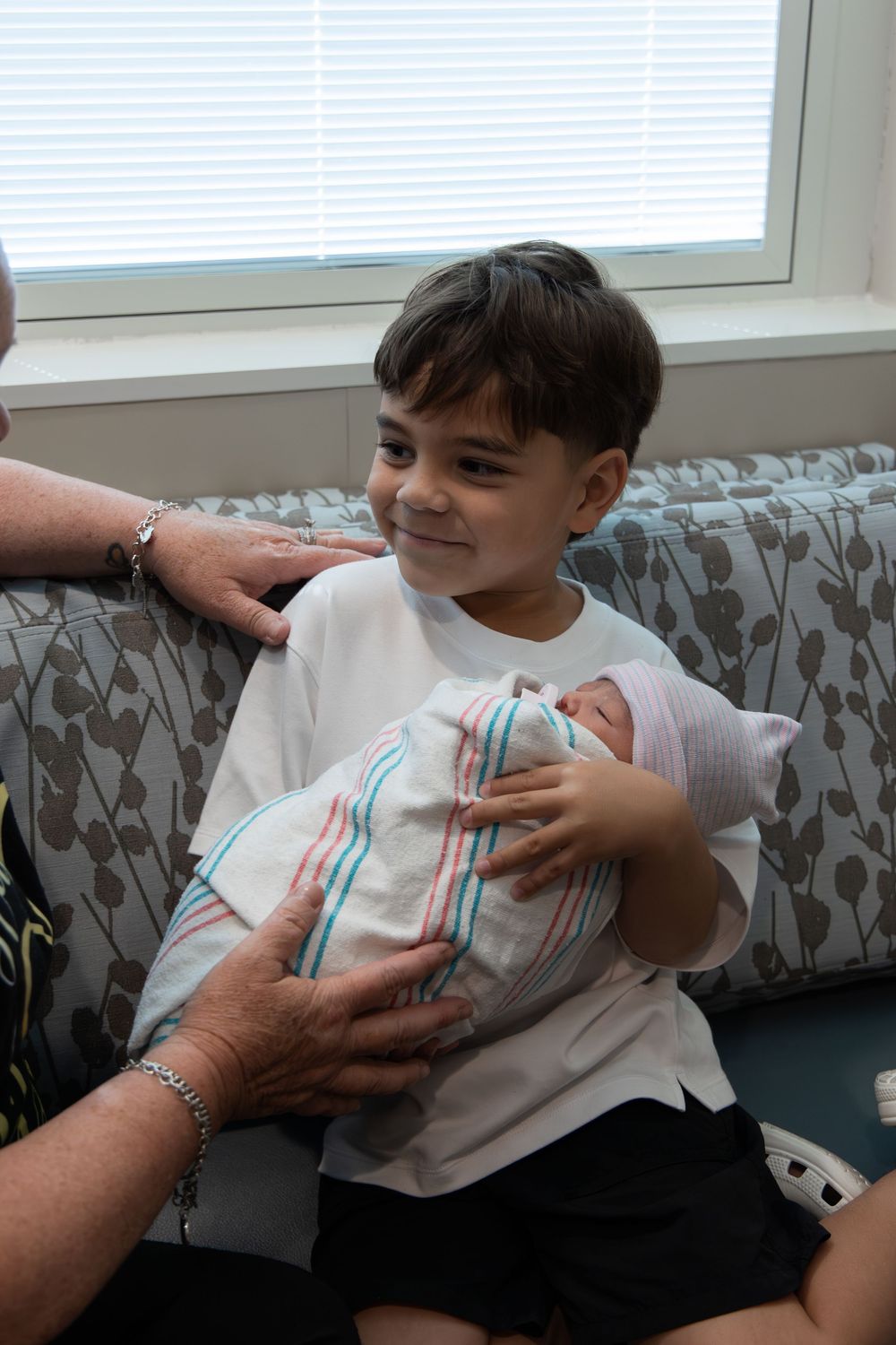 Young boy smiling while carefully holding a swaddled newborn baby on a gray floral couch.