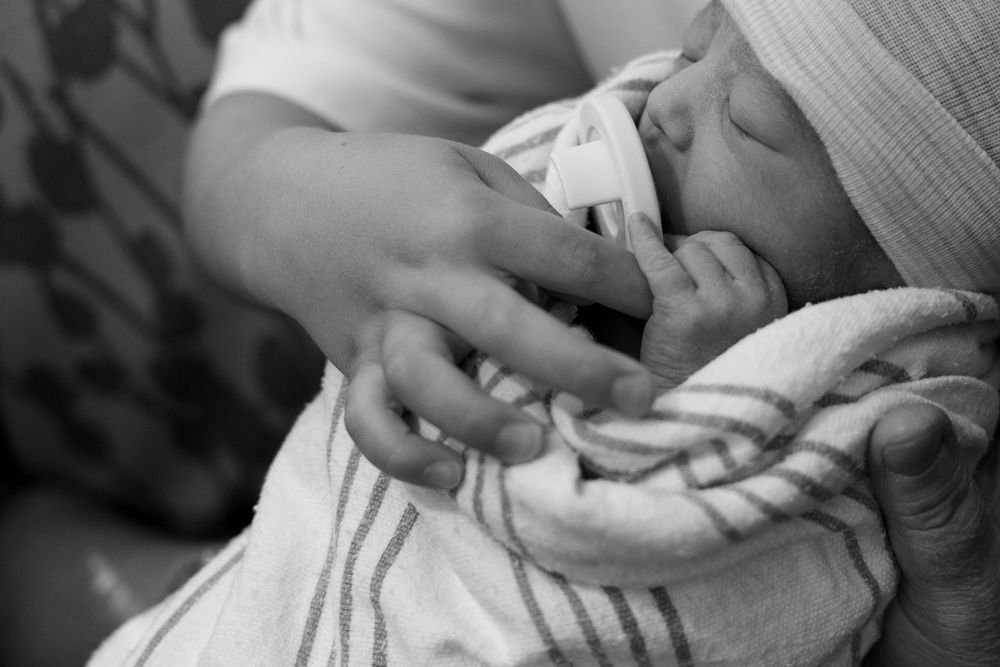 Black and white photo of a newborn baby swaddled in a striped blanket, held gently by an adult's hands.