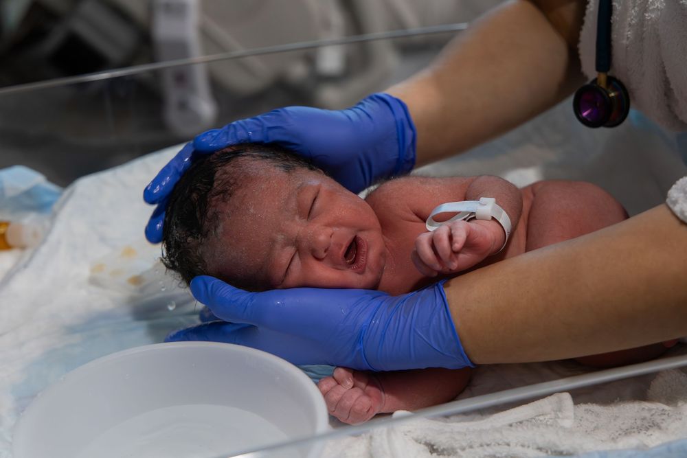 Newborn baby being held by medical professional wearing blue gloves in hospital delivery room.