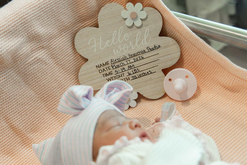 Newborn baby sleeping beside a wooden Hello World birth announcement sign with name, date, time, and weight.