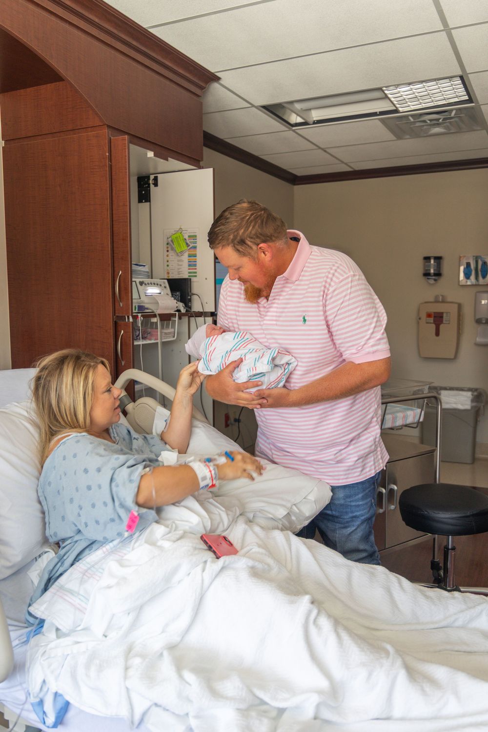 New parents admire their newborn baby in a hospital room, with mother resting in bed and father holding the infant.