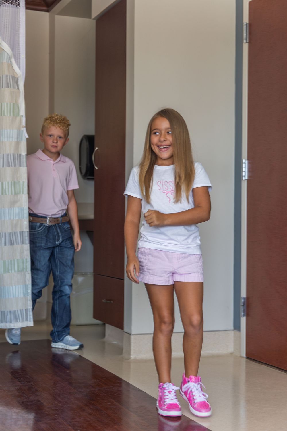 Two young children, a smiling girl in pink shorts and a boy in a pink polo, stand in a hospital room doorway.