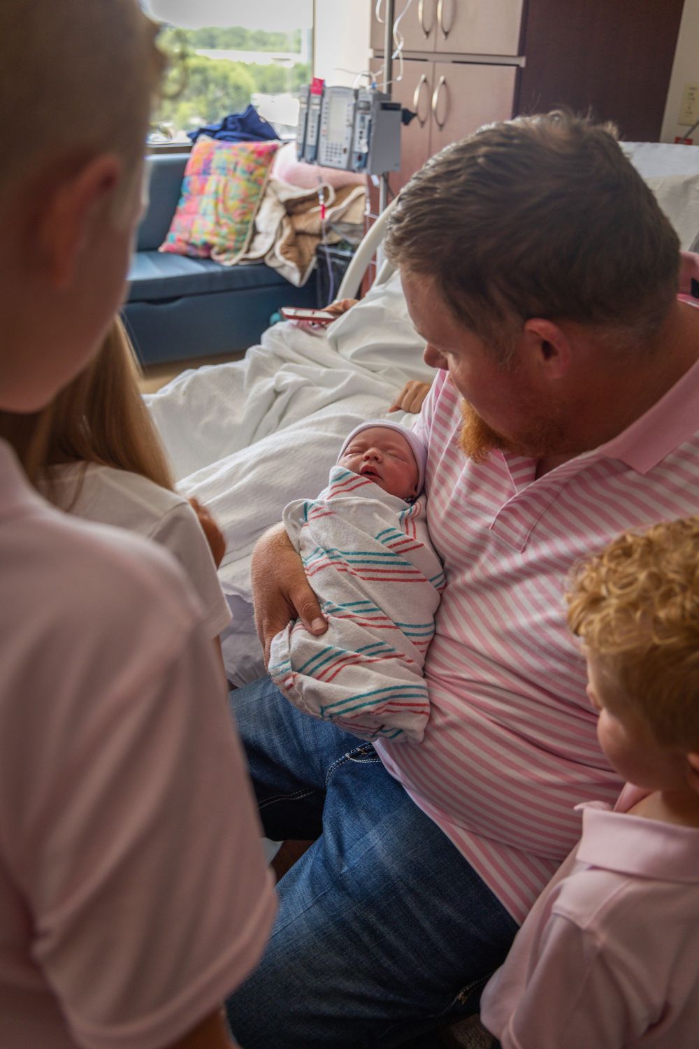 Family gathered in hospital room admiring newborn baby held by grandfather, with curly-haired child nearby.