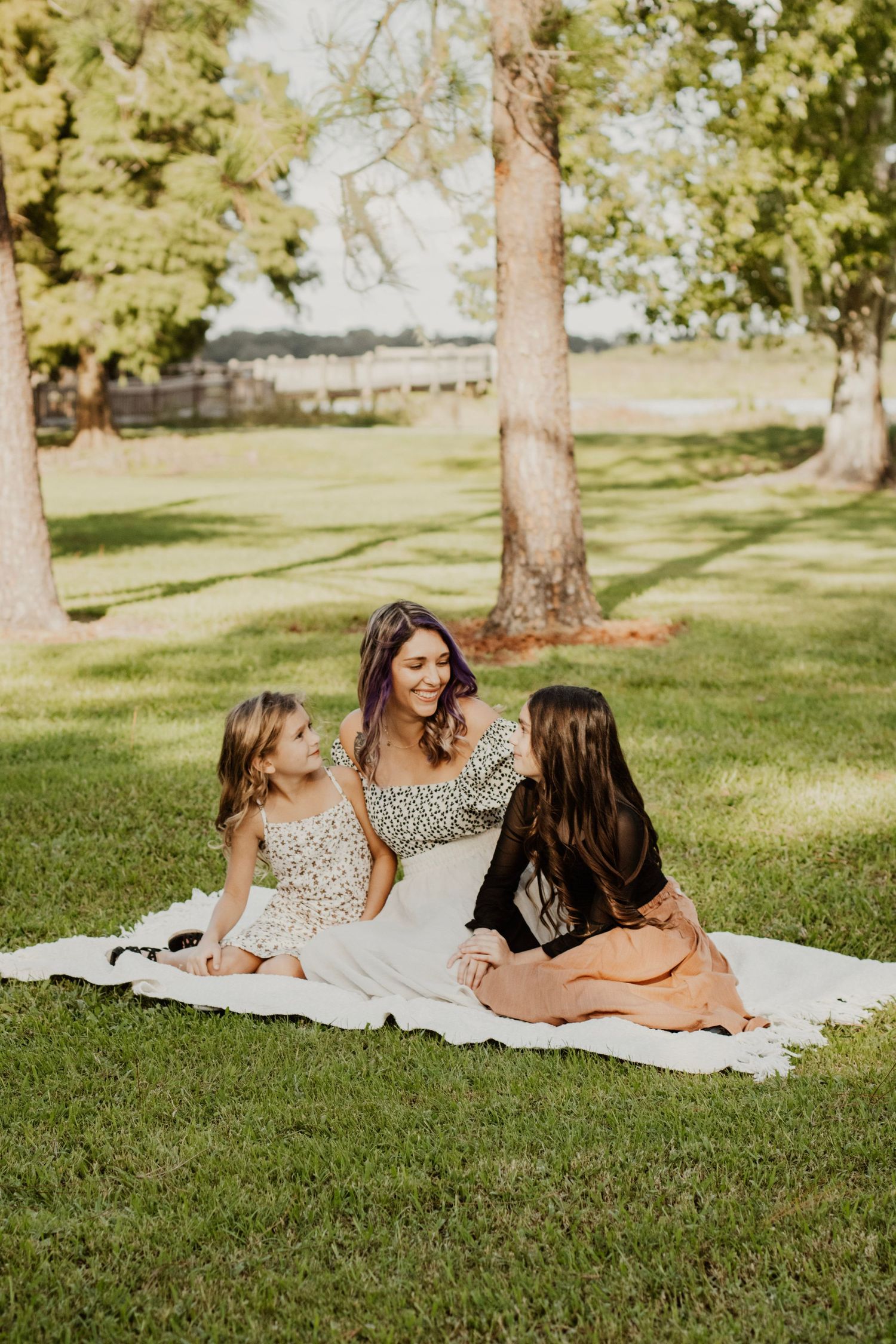 A mother and two daughters sitting together on a blanket in a lush green park, smiling warmly.