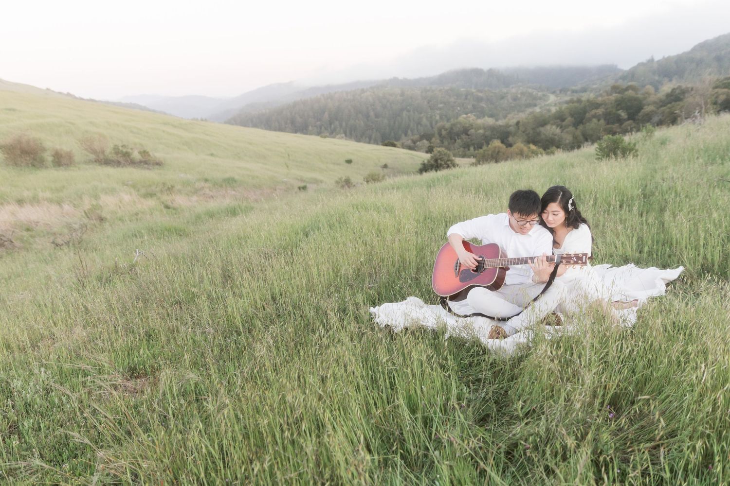 Couple sitting in green meadow playing acoustic guitar with rolling hills and forest in background.