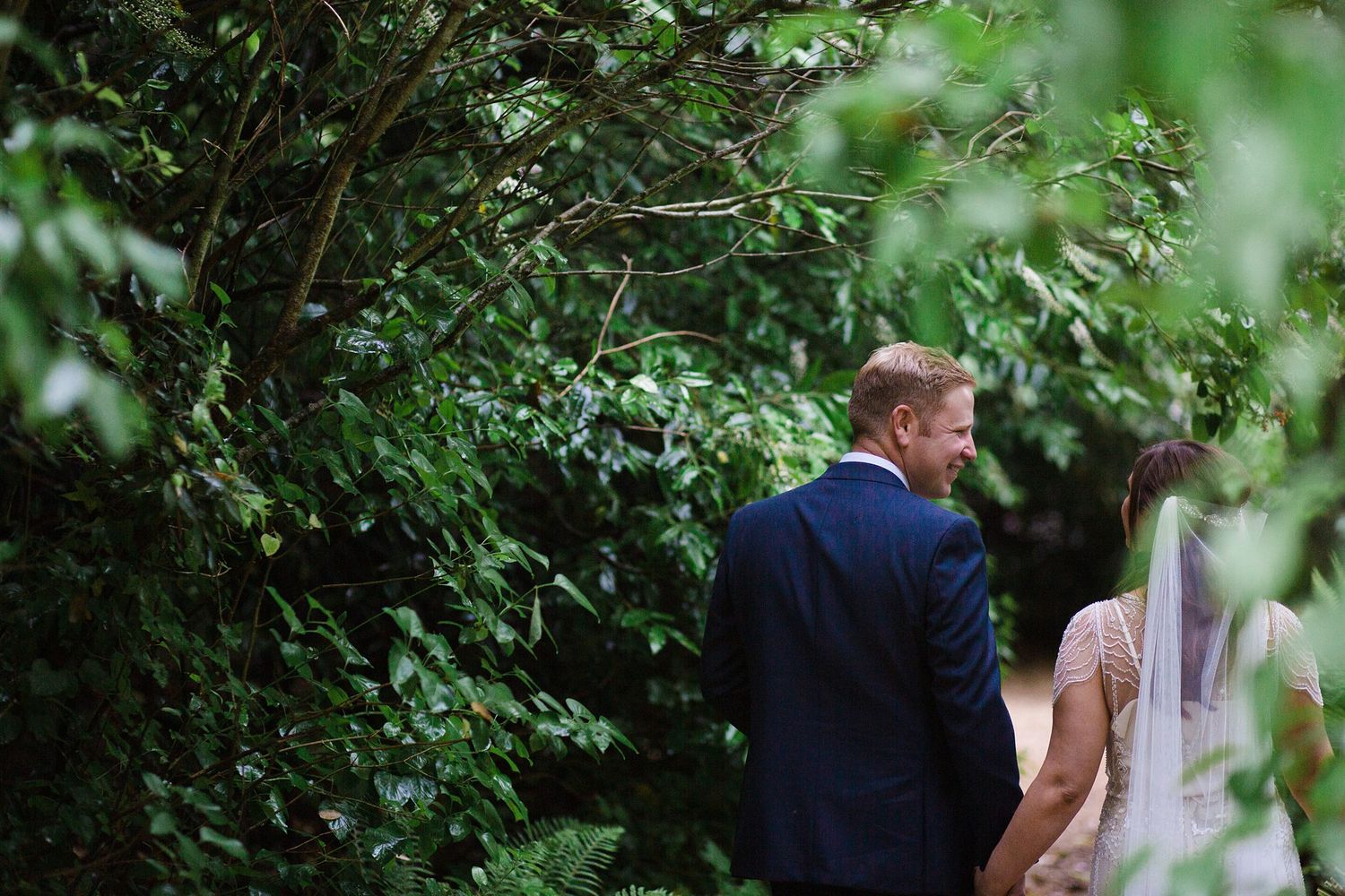 A bride and groom walk hand in hand through a lush green garden pathway on their wedding day at the Tangled garden, GrandPre.