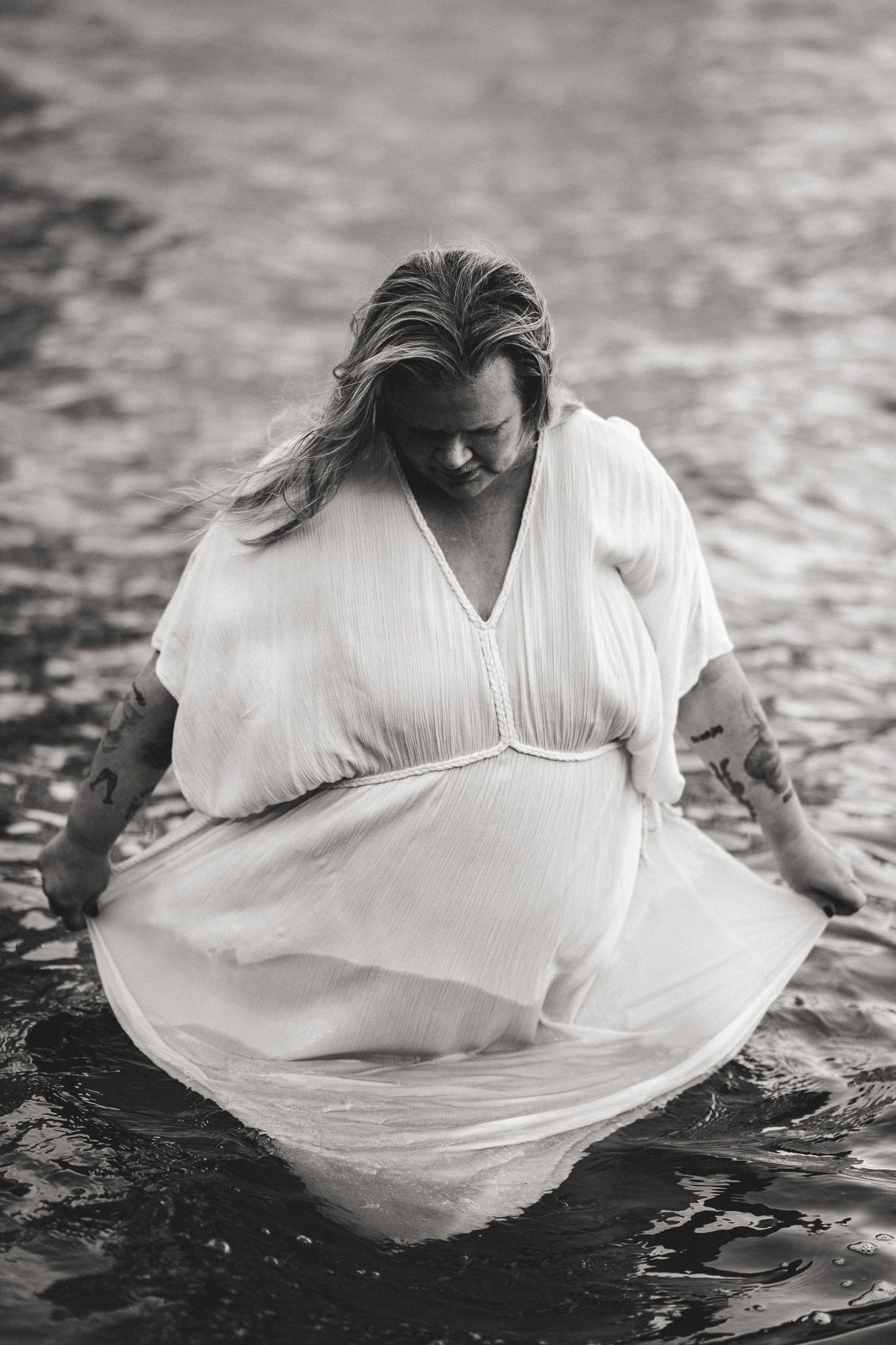 Svartvitt porträttfoto av Fotograf Jessica Lindqvist - en kvinna som håller i sin klänning stående i vattnet.