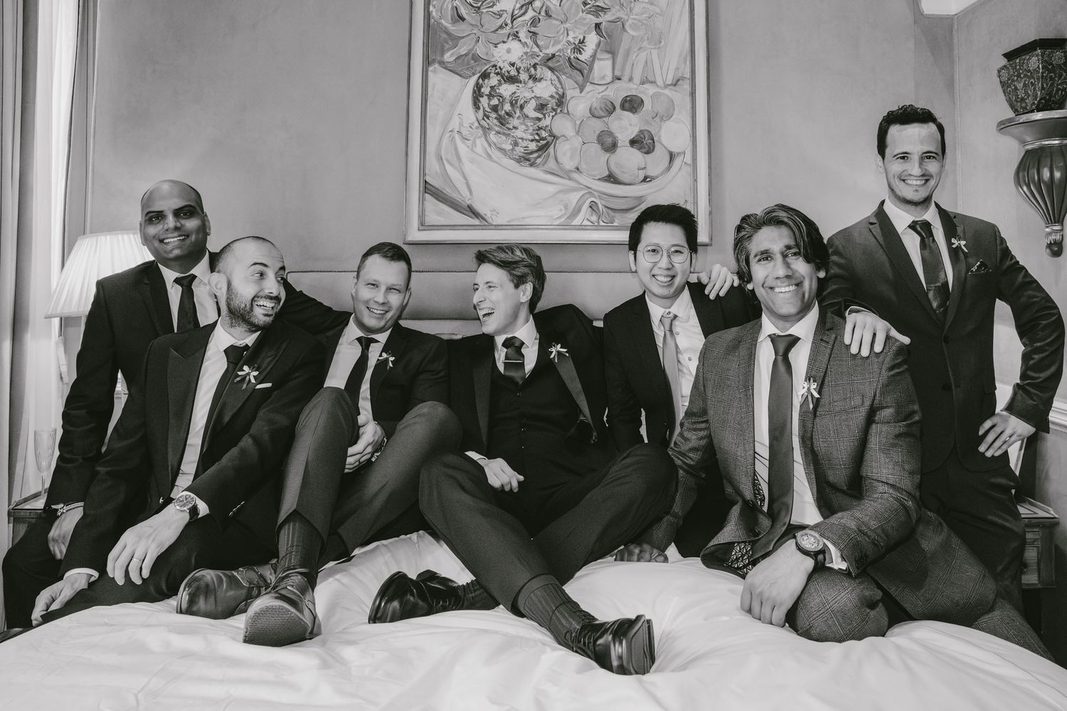 Black and white photo of seven groomsmen in suits laughing and posing together on a bed before a wedding.