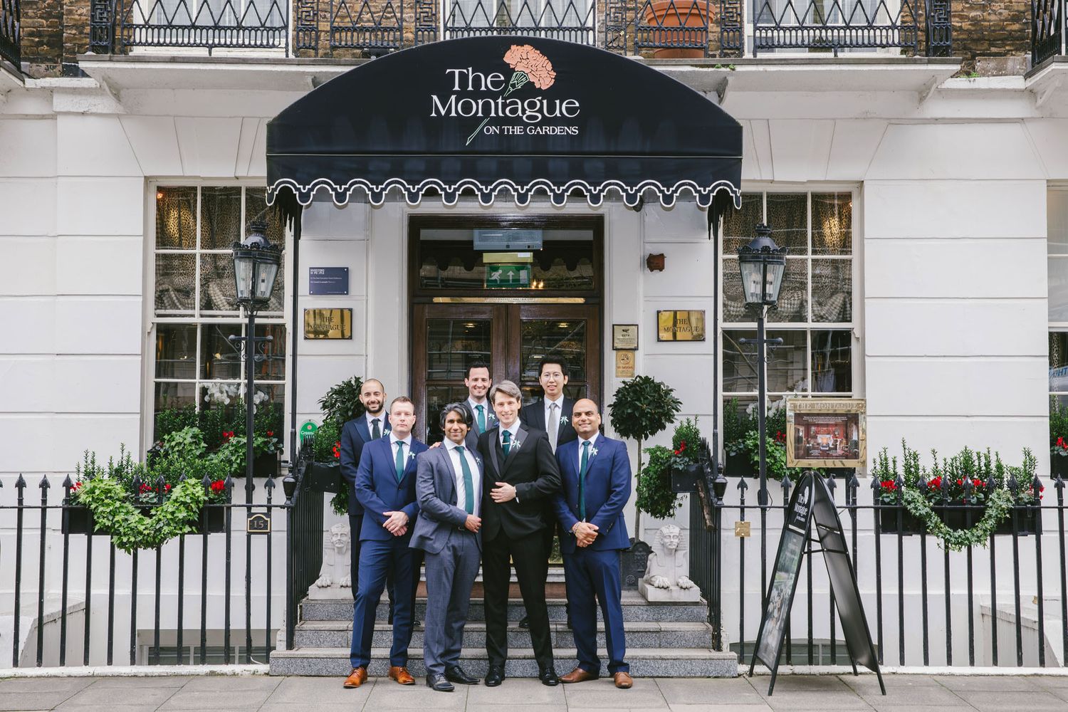Group of men in suits posing outside The Montague on the Gardens hotel entrance in London.