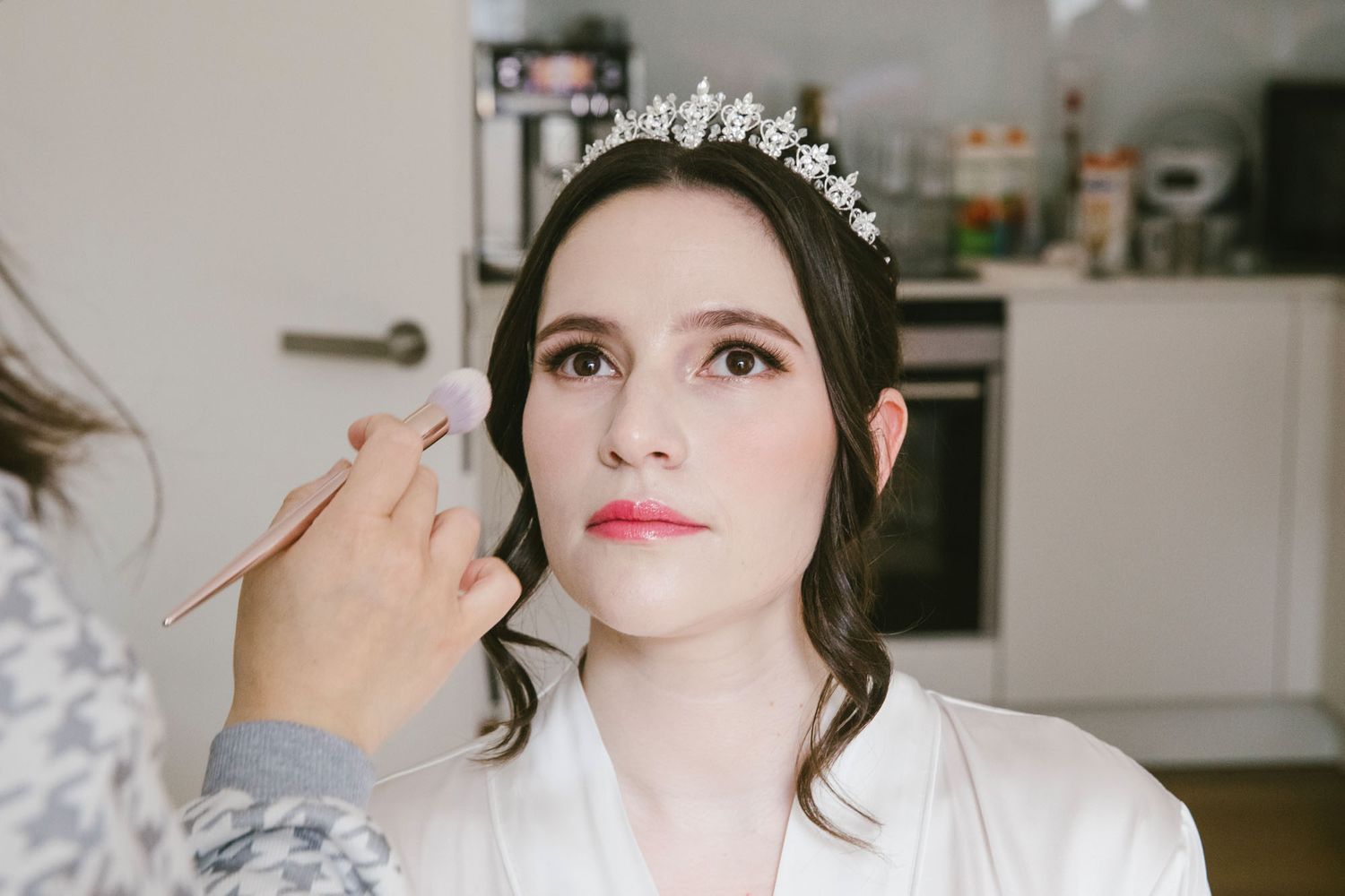Bride in white robe wearing floral tiara gets makeup applied by artist before wedding ceremony.