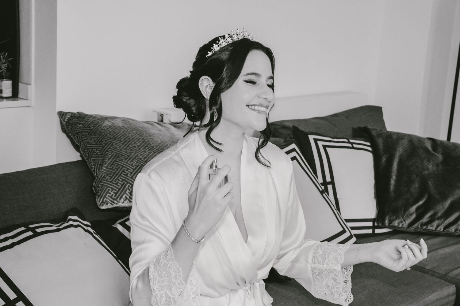 Bride in white lace robe wearing tiara sits on sofa smiling, black and white wedding preparation photo.