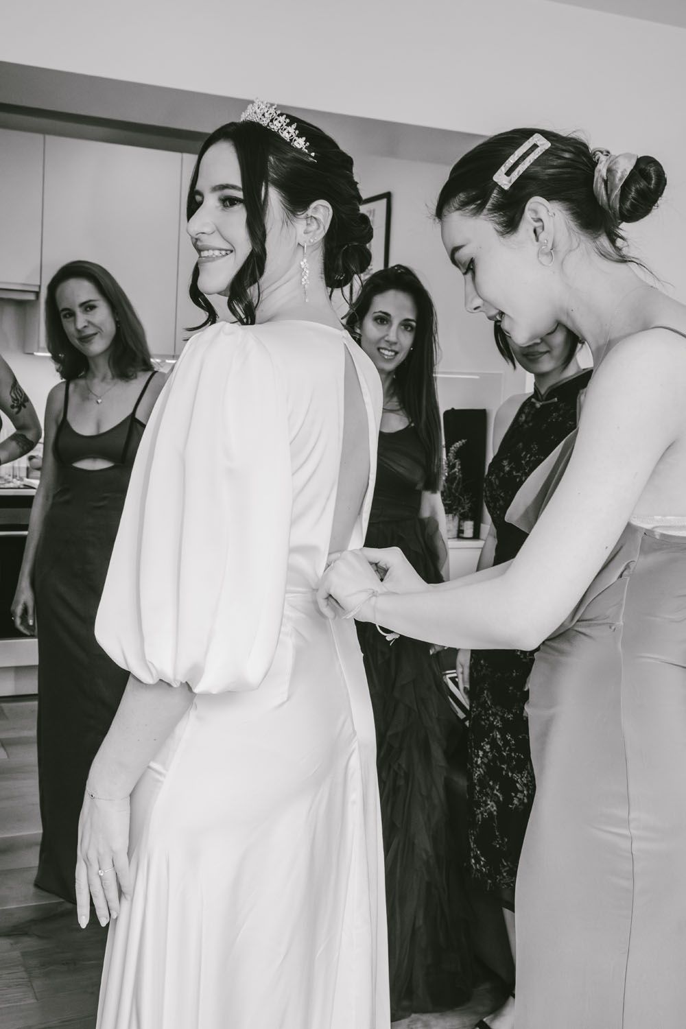 Bridesmaids helping bride fasten her white wedding dress in black and white photo during bridal prep.