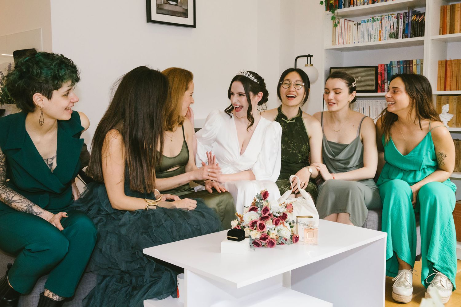 Bride in white robe laughing with bridesmaids in green dresses during bridal party getting ready celebration.