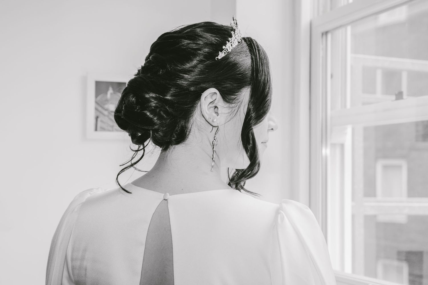 Black and white photo of a bride with elegant updo hairstyle and tiara, wearing a white dress, standing by a window.