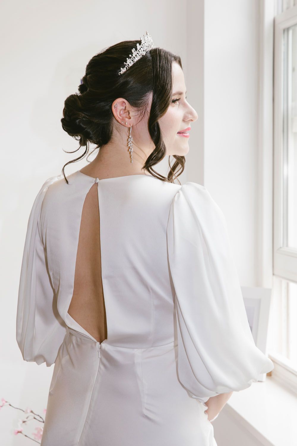 Bride in elegant white open-back wedding gown with puff sleeves and crystal tiara gazing out window.