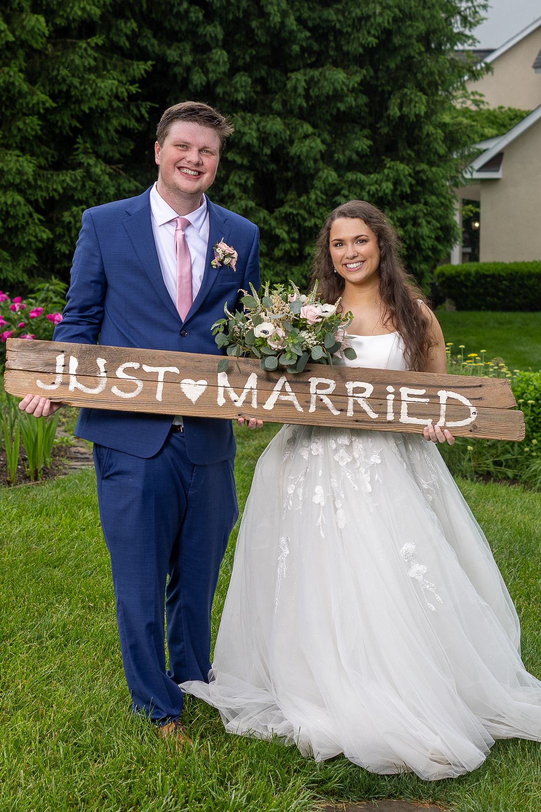 Bride in white gown with long veil stands before large windows in rustic wood venue, smiling over her shoulder.