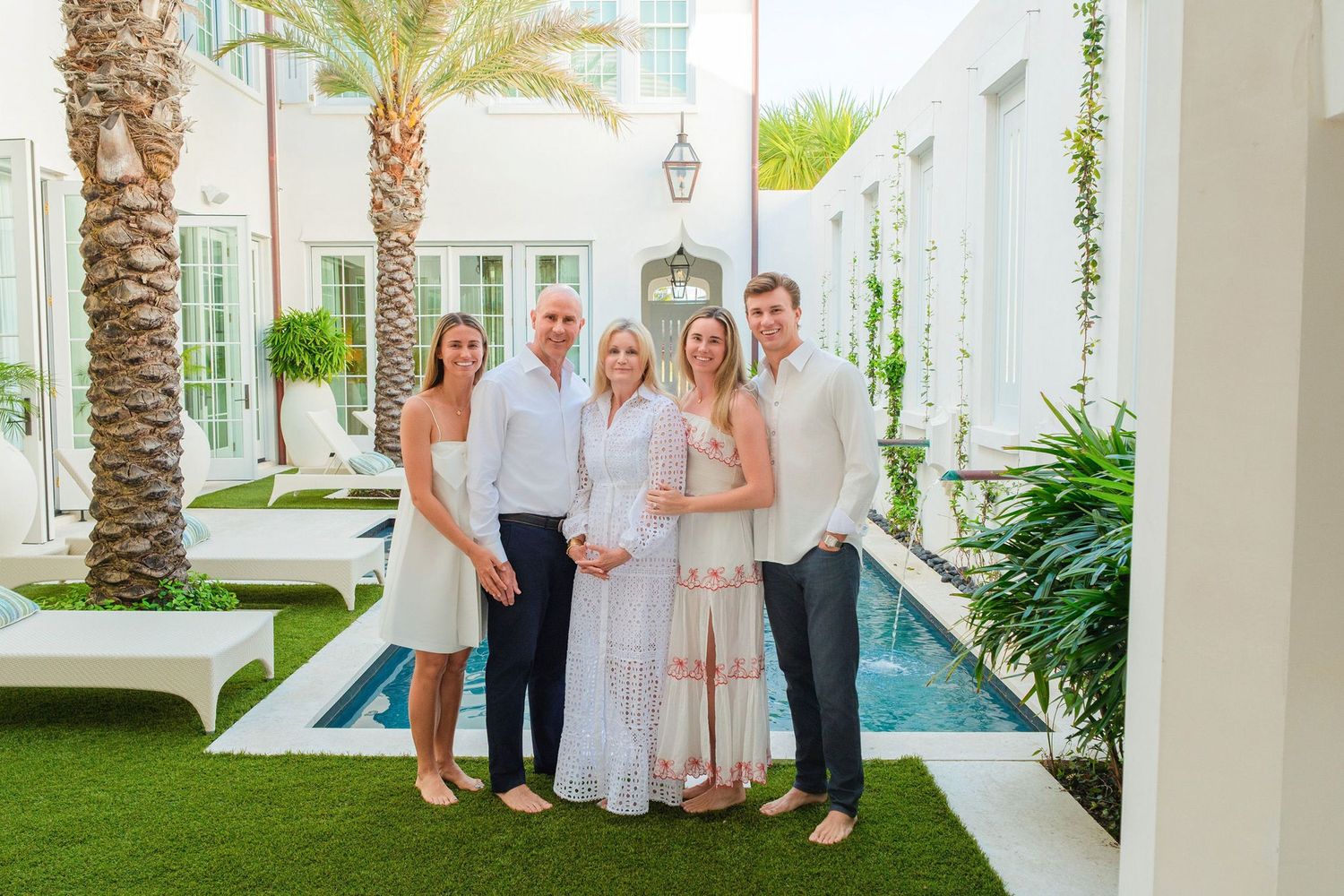 Five people posing together in an elegant outdoor courtyard with a pool, lush greenery, and white architecture.