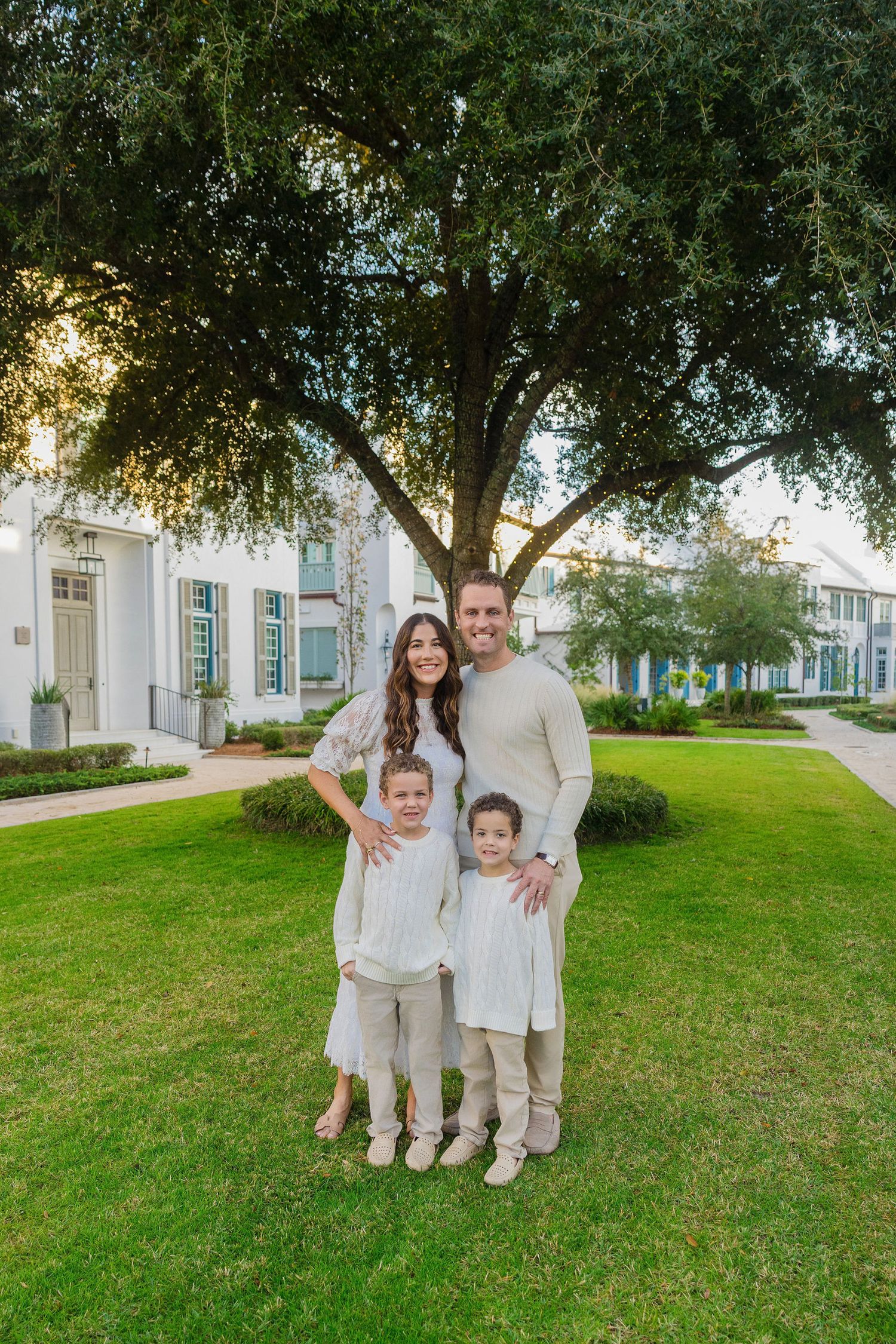 Family of four in neutral outfits posing on green lawn under large oak tree beside white colonial-style buildings.