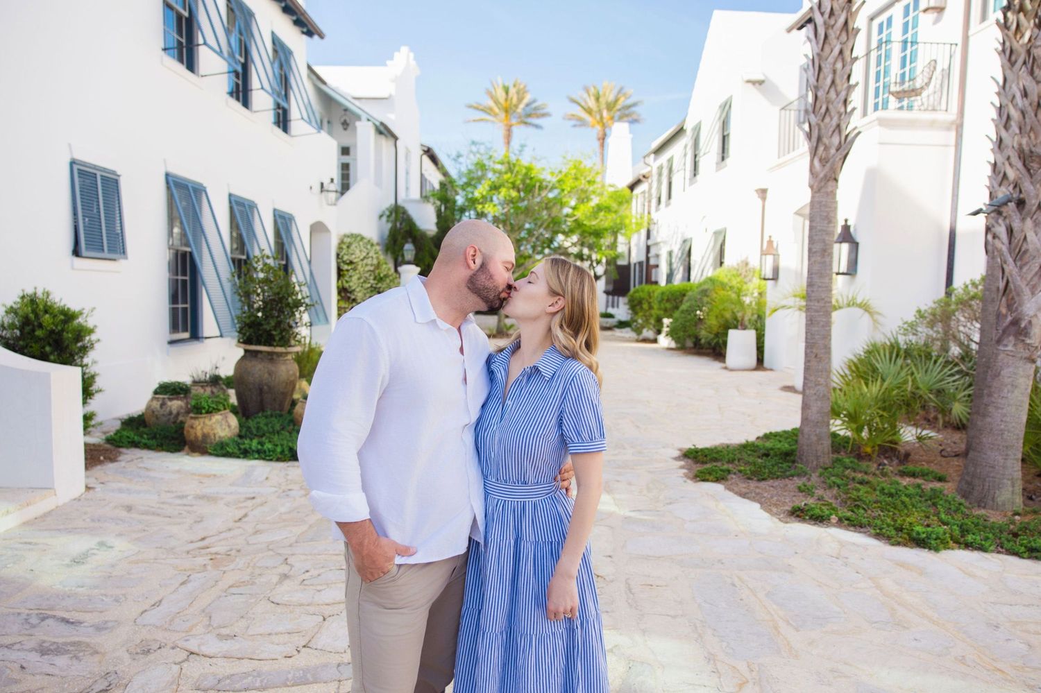 Couple sharing a romantic moment kissing in a charming white Mediterranean-style courtyard with palm trees.