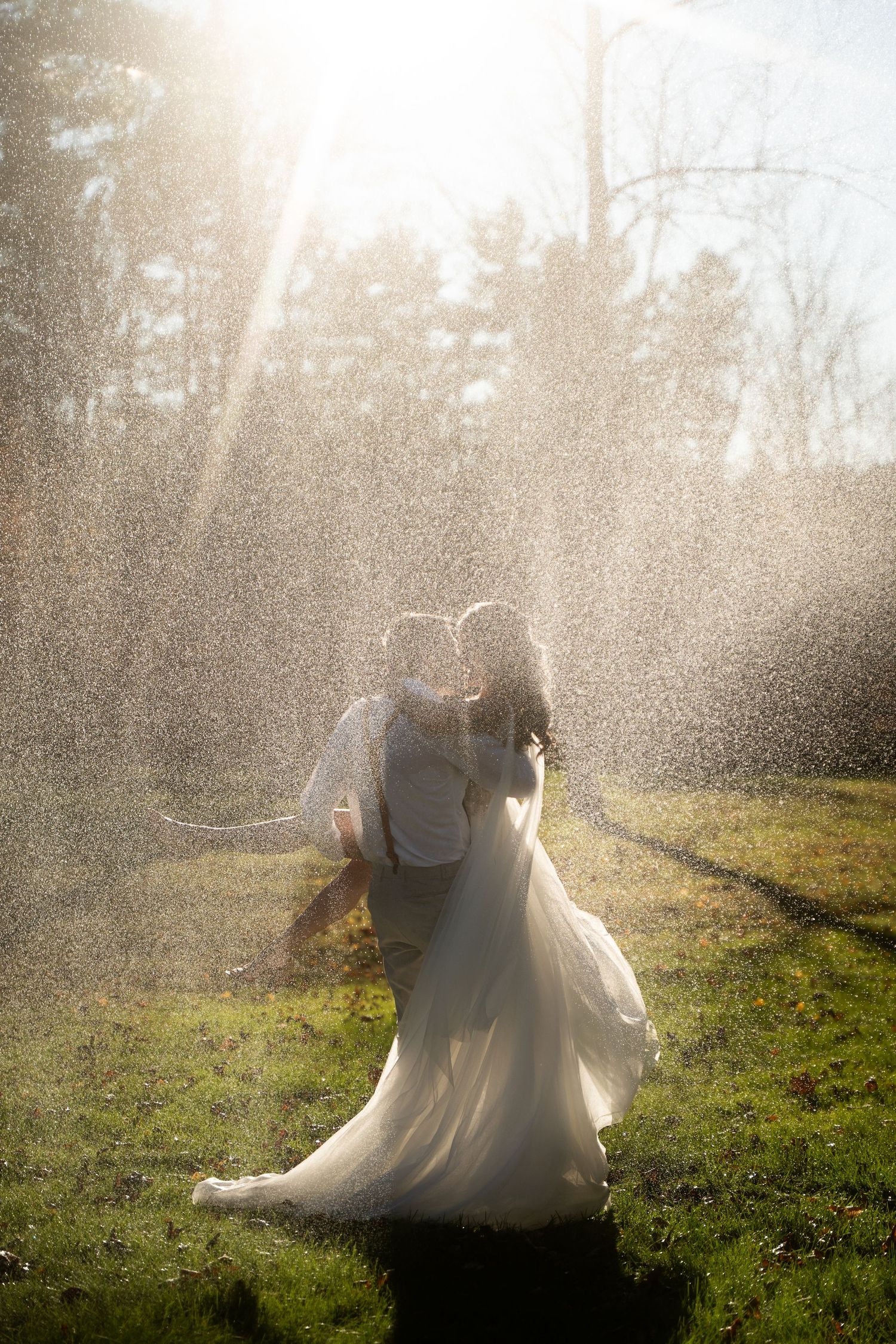 Bride and groom kissing in a magical spray of water mist, backlit by golden sunlight in a lush green garden. Westerly Rhode Island