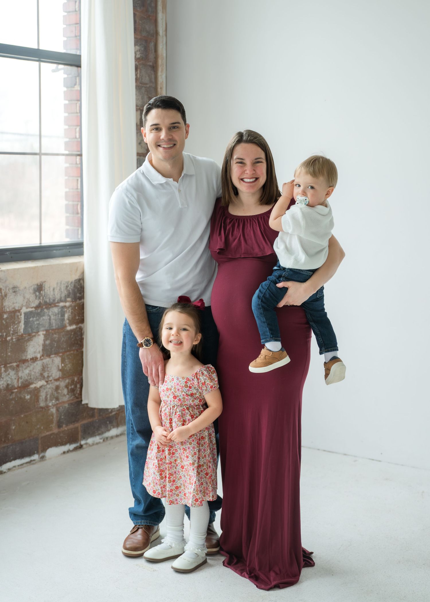 Smiling family of four posing together in a bright studio, mom in burgundy maternity gown holding toddler son.
