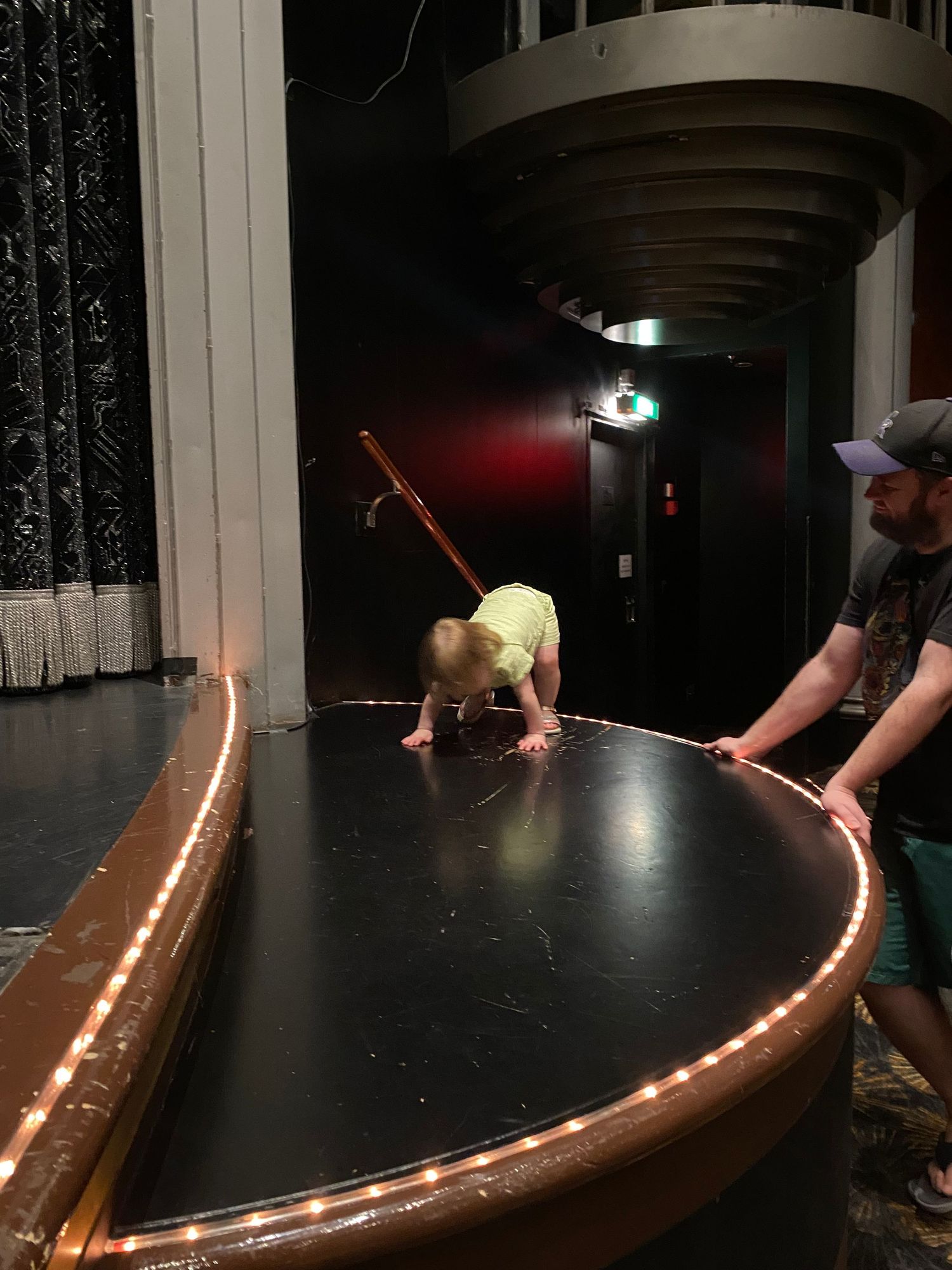 A baby crawls on a theater stage near footlights while an adult watches nearby.