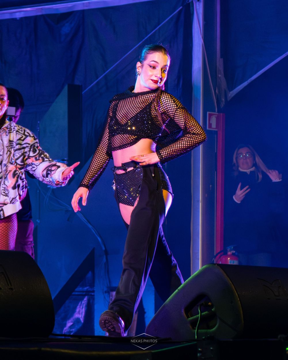 Female dancer performing on stage in black sequined outfit and mesh top under blue and purple stage lights.